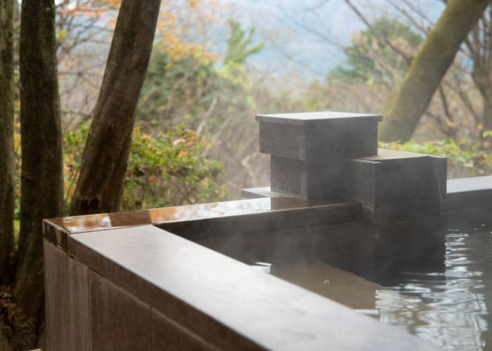 The corner of a wooden Japanese onsen bath, looking out into the trees from the steaming water.